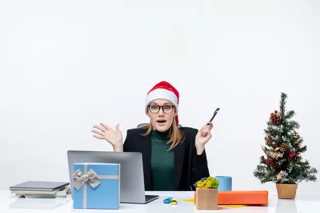 Shocked blonde woman with a santa claus hat sitting at a table with a christmas tree and a gift on it in the office on white background Free Photo